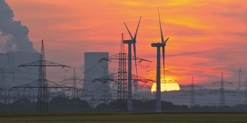 Das Bild zeigt gegen die untergehende Sonne zwei Windkraftanlagen, einen Kühlturm und mehrere Strommasten.
