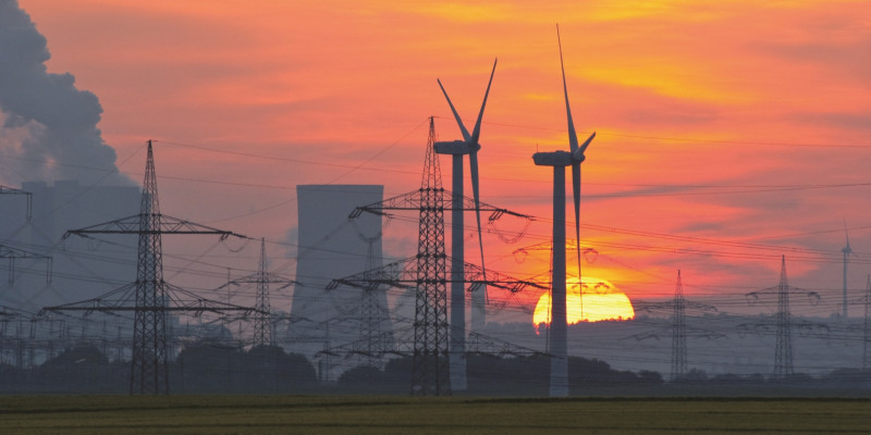 The picture shows two wind turbines, a cooling tower and several electricity pylons against the setting sun.