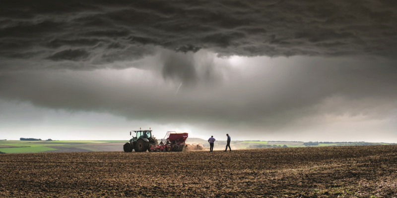 Das Bild zeigt im Vordergrund einen Acker. Ein Traktor sät gerade und zwei Männer kontrollieren die Einsaat. Im Hintergrund ist eine weiter landwirtschaftlich genutzte Landschaft zu sehen. Der Himmel ist sehr dunkel, es naht ein Gewitter.