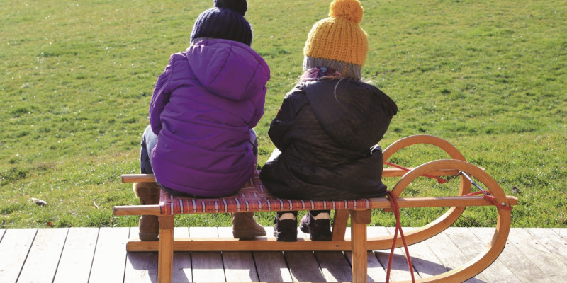 Das Bild zeigt zwei Kinder von hinten, die mit Pudelmützen und Winterjacken nebeneinander auf einem Schlitten sitzen, der auf einer Holzterrasse vor einer grünen Wiese steht.