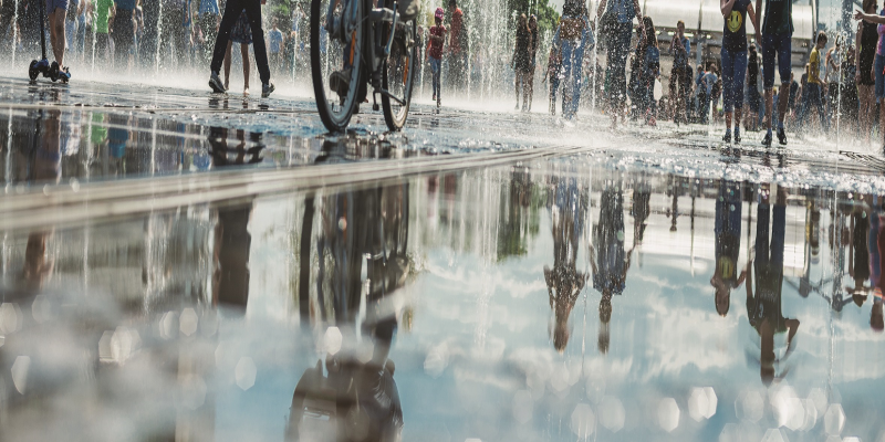Das Bild zeigt eine Wasserfläche in eine Stadt, offensichtlich ein Brunnen. Es laufen mehrere Menschen über die Wasserfläche und spiegeln sich darin.