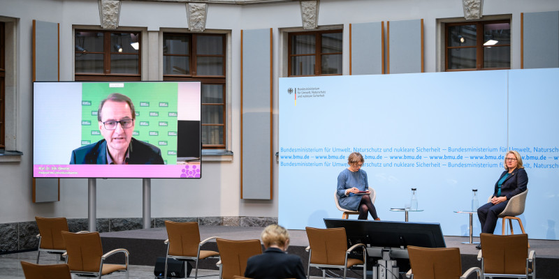 Der Blick geht in dem hellen historischen Innenhof von hinten über das Publikum hin zum Podium vor blauem Hintergrund, wo die Umweltministerin und eine Konferenzteilnehmerin sitzen im Gespräch mit dem Präsidenten des Umweltbundesamtes, der links des Podiums auf dem großen flachen Bildschirm zugeschaltet wurde.