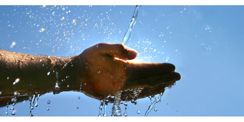 Zwei Hände werden in einen Wasserstrahl gehalten. Im Hintergrund scheint die Sonne