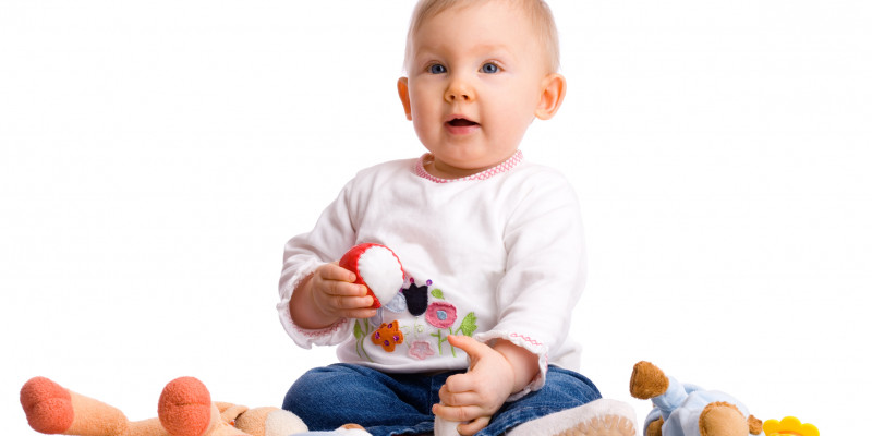 Ein Baby sitzt neben Kuscheltieren, es hält einen Ball in der Hand und guckt erwartungsvoll nach oben.