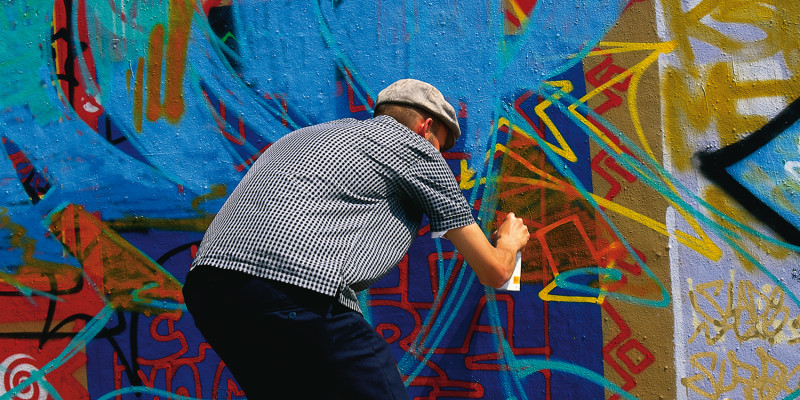 Mann der gebeugt vor einer Wand steht und ein bereits existierendes Graffitti übersprüht