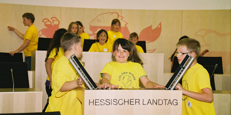 Drei Kinder in einem gelben T-Shirt stehen hinter und neben einem Pult auf dem Hessischen Landtag