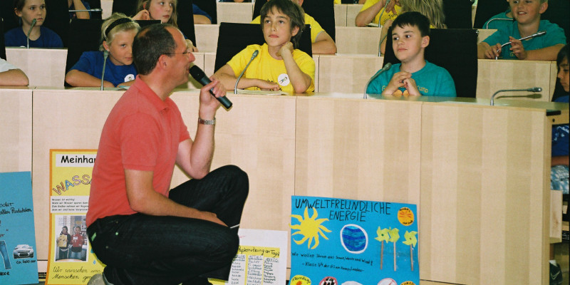Ein Mann mit Mikrofon in der Hand hockt vor Kindern fürs Interview.