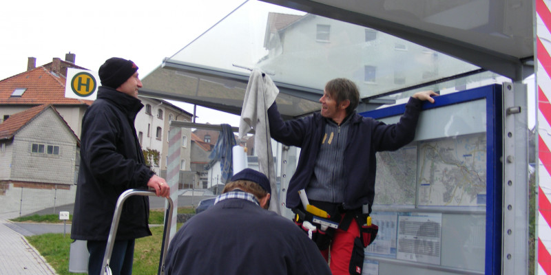 Personen stehen an einer überdachten Bushaltestelle und bekleben das Dach mit der Folie
