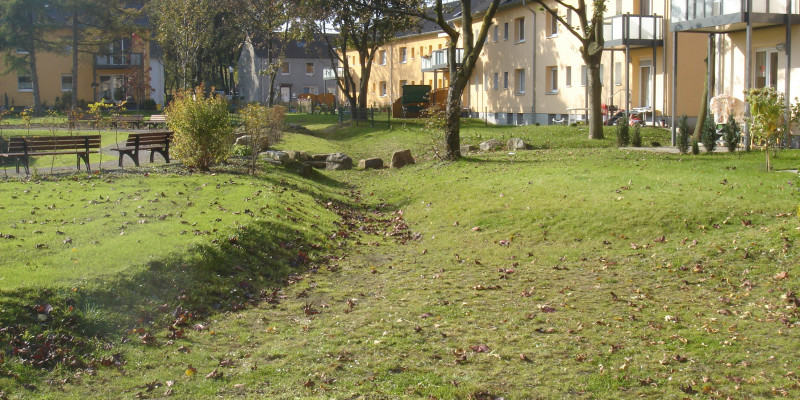 Auf dem Rasenboden verlaufen Rinnen für Regenwasserabfluss. Im Hintergrund stehen Wohnhäuser mit Balkonen und Terassen zum Rasen hin.