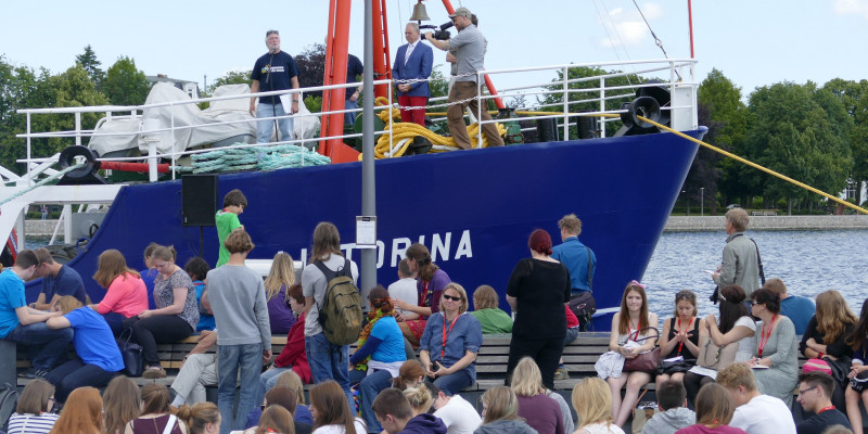 Menschenmenge vor einem Schiff, auf dem Redner und Kameramann stehen.