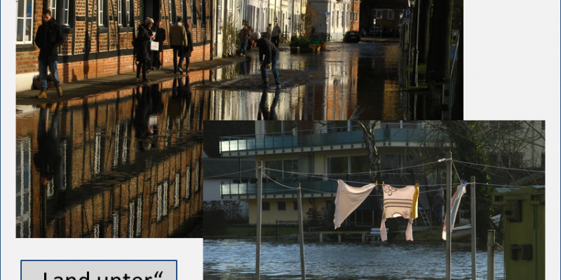 Wasser überflutet die Straße in der historischen Altstadt. Am linken Bildrand bewegen sich Menschen auf dem verbliebenen schmalen Bürgersteig vor den Altstadtfassaden. Auf der rechten Seite säumen Bäume und Laternen die Straße. Im Vordergrund rechts sieht man zum Trocknen aufgehängte Wäsche auf einem Wäscheständer tief im Wasser stehend, im Hintergrund befindet sich ein modernes Wohnhaus.