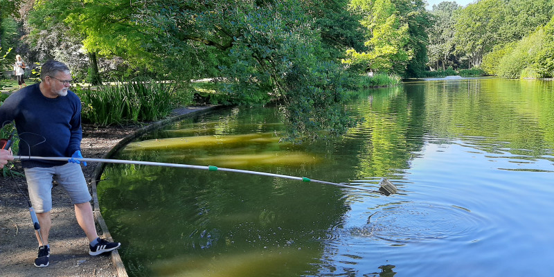 Gewässeruntersuchung am Waller Park See Bremen: ein Mann steht mit einer langen Stange am Wasser vor grünbelaubten Bäumen.