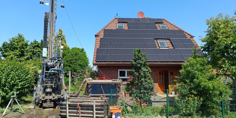 Ein geklinkertes Haus steht quer zur Straße, das Dach ist mit Photovoltaik belegt. Vor dem Haus im Vorgarten steht ein hohes Bohrgerät, daneben Stangen auf einer Palette und ein Container.