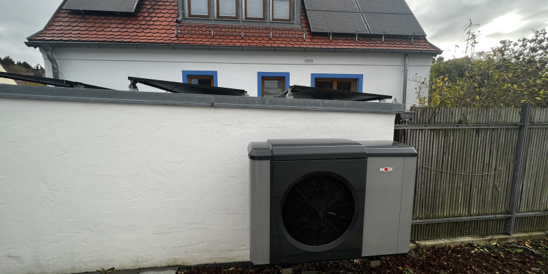 Eine Wärmepumpe an einer Grundstücksmauer, im Hintergrund das Einfamilienhaus.