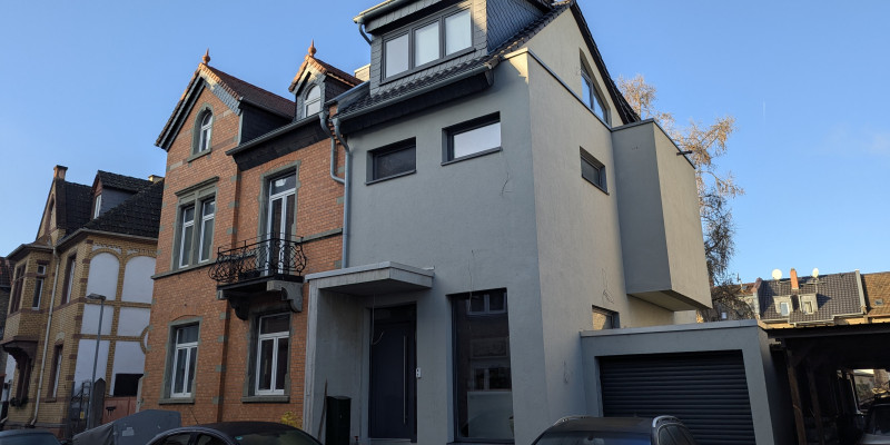 Die Fassade eines zweigeschossigen, rot geklinkerten Altbaus mit einem modernen, grau verputzten Anbau.