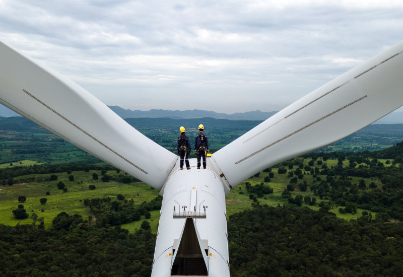 Zwei Menschen i9n Arbeitskleidung stehen auf einer Winkraftanlage und schauen in einer bewaldete Landschaft.