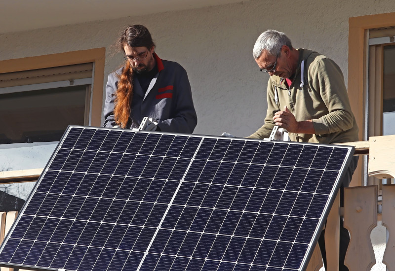 Zwei Personen installieren ein Solarpanel an Holzgeländer eines Gebäudes, im Hintergrund sind Fenster zu sehen.