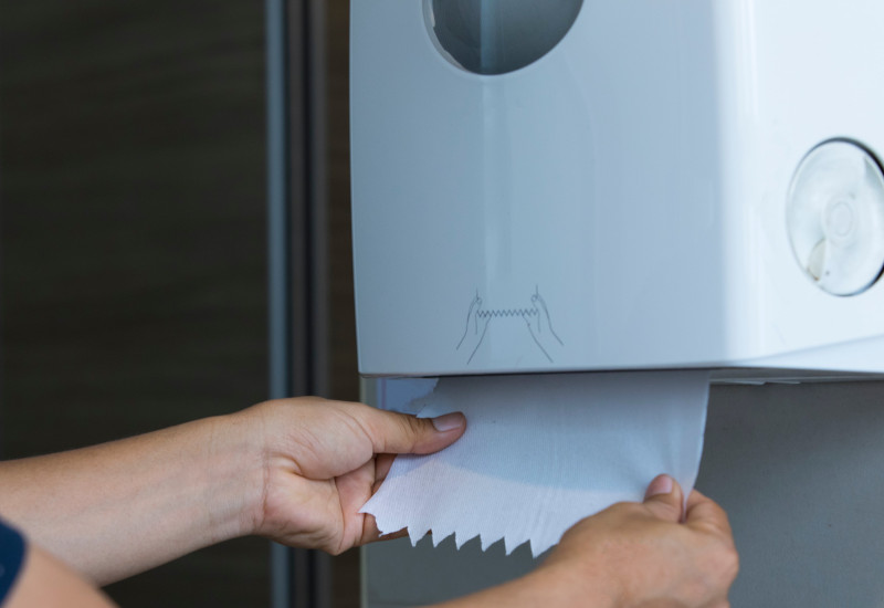  Eine Person zieht mit beiden Händen ein weißes Papierhandtuch aus einem an der Wand befestigten Spender.