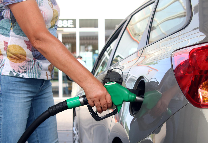  Eine Person in Jeans und einem T-Shirt mit Blumenmuster hält eine grüne Zapfpistole in den Tank eines silberfarbenen Autos.