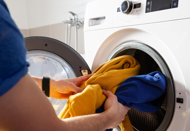 Eine Person steckt bunte Wäsche in eine Waschmaschine. Die offene Tür der weißen Waschmaschine ist zu sehen. Im Vordergrund liegt ein Haufen bunter Kleidung.