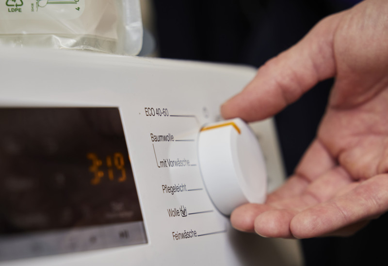 Eine Nahaufnahme zeigt eine Hand, die den Drehknopf einer Waschmaschine auf ein Programm einstellt. Auf dem Bedienfeld der Maschine sind verschiedene Waschprogramme wie „ECO 40-60“, „Baumwolle“ und „Pflegeleicht“ zu sehen.