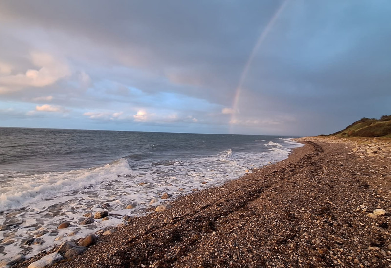 Wellen der Ostsee schlagen an einen Steinstrand, der in grüne Hügel übergeht. Der Himmel ist über den Hügeln graublau aber über dem Meer hellblau mit weißen Wolken. Ein Regenbogen beginnt über dem Meer.  