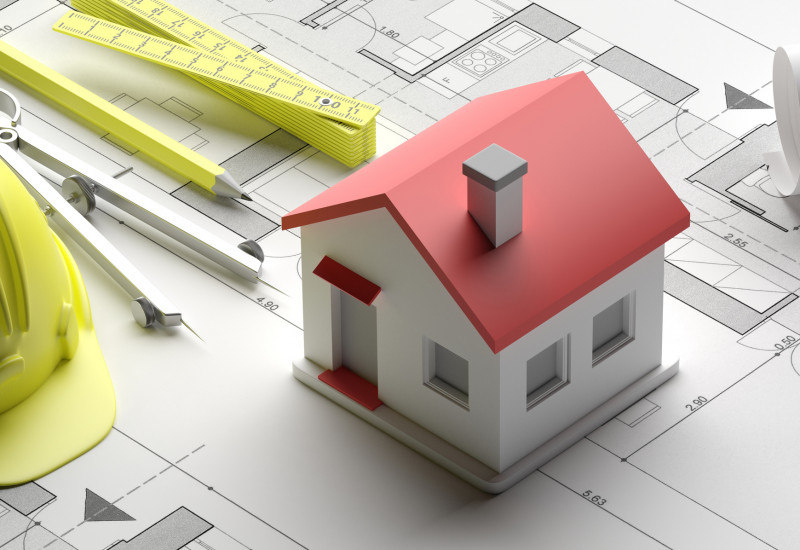 Das Bild zeigt einen Bauplan und ein Modell für ein Eigenheim sowie Zirkel, Bleistift, Zollstock und Schutzhelm.