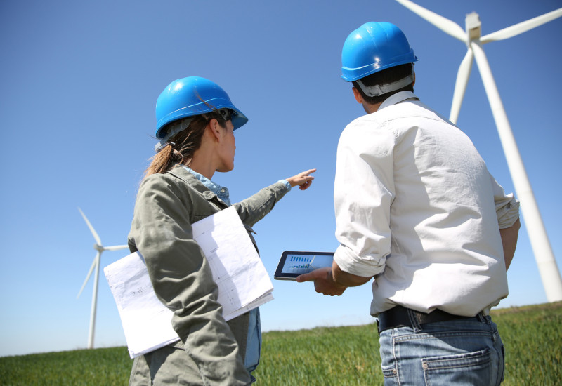 Das Bild zeigt im Vordergrund zwei Ingenieure, welche sich über Pläne beugen. Im Hintergrund sind Windenergieanlagen zu sehen. 