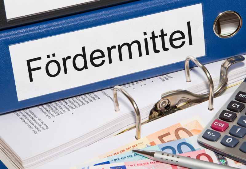 Foto: Aktenordner mit Beschriftung „Fördermittel“, davor auf einem Tisch liegen Geld, Taschenrechner und Kugelschreiber.
