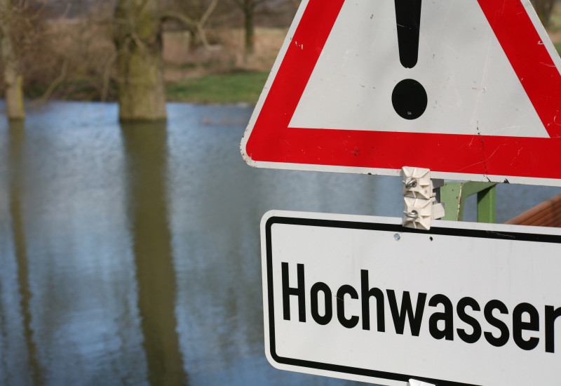 Foto: Ein überschwemmtes Gebiet während eines Hochwasserereignisses. Im Vordergrund steht ein „Achtung Hochwasser“ Schild