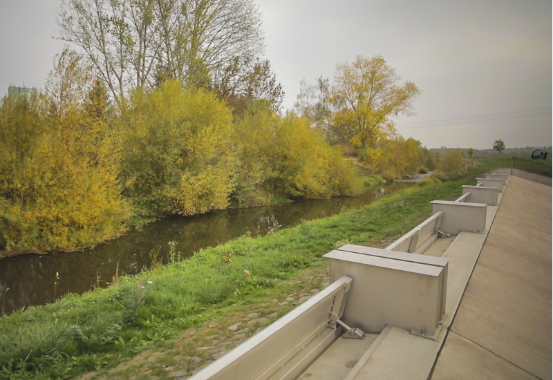 Foto: Ein Klappenwehr aus Beton und Stahl entlang der Helme. Das Ufer im Bereich vor dem Klappenwehr ist mit Gräsern bedeckt. Die andere Uferseite ist naturnah mit Gehölzen bestanden.