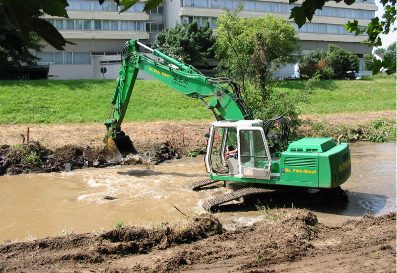 Foto: Ein Bagger bei der Bearbeitung der Ufer eines Fließgewässers. Am Ufer sind einzelne Bäume zu sehen. Im Hintergrund eine Brücke.