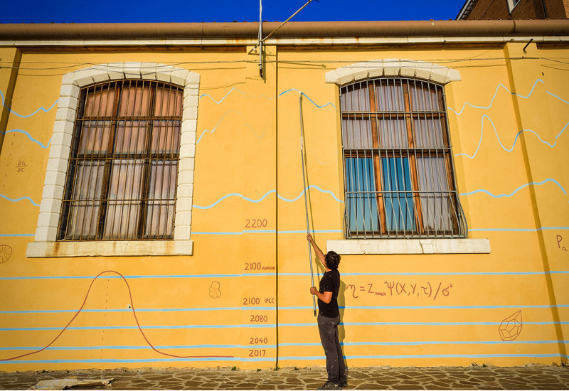Das Foto zeigt den Künstler Andreco bei der Bemalung einer Hauswand in Venedig. Die Hauswand trägt bereits einige fiktive Hochwassermarken bis 2100. Das Bild visualisiert das Risiko des globalen Meeresspiegelanstiegs für den Menschen jenseits einer globalen Erwärmung auf 1,5 °C. 