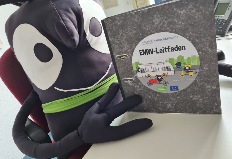 Stoffpuppe des EMW-Maskottchens hält den neuen EMW-Leitfadenordner hoch.