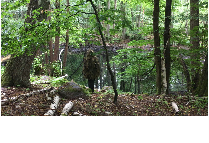 Fotografie eines Menschen, der mit Naturmaterialien verkleidet ist und im Wald vor einem Teich steht