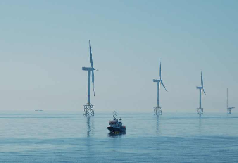 Das Bild zeigt im Meer stehende Windräder eines Offshore-Windparks und ein dazwischen schwimmendes Schiff.