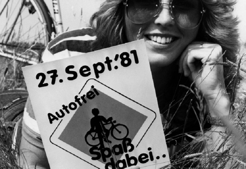 eine Frau lmit Sonnenbrille iegt neben ihrem Fahrrad auf einer Wiese und hält ein Schild in die Kamera: 27. Sept.´81, Autofrei – Spaß dabei“