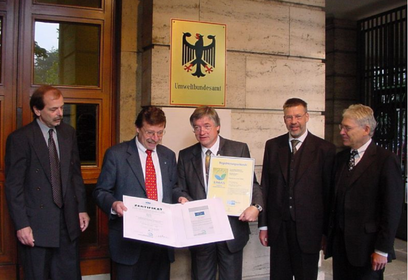Gruppenfoto: fünf Männer im Anzug vor einem Gebäudes des Umweltbundesamtes, zwei halten ein Zertifikat und eine Urkunde in die Kamera