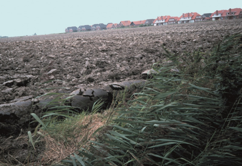 ein kahler, umgepflügter Acker, im Hintergrund ein Neubaugebiet