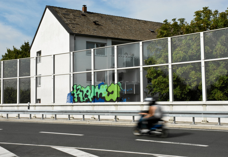 ein Wohnhaus liegt direkt an einer Stadtautobahn hinter einer Lärmschutzwand, gerade fährt ein Motorrad vorbei