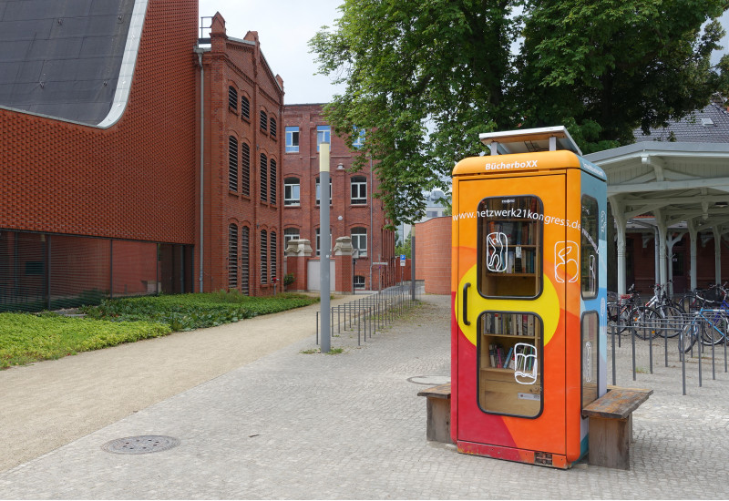 eine bunt angemalte Telefonzelle ist innen zu einer Miniblibliothek ausgebaut, auf dem Dach ein Solarmodul, außen eine Holzbank