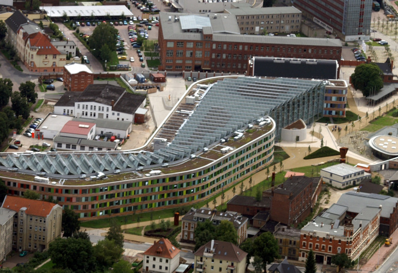 das UBA-Gebäude in Dessau-Roßlau von oben fotografiert, das Gebäude ist langgestreckt, hat eine Fassade aus Holt und bunten Glasplatten und schräggestellte Solarzellen auf dem gesamten Flachdach