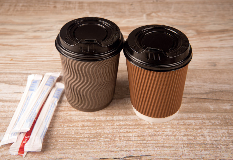 zwei „Coffee to go“-Becher auf einem Tisch, daneben zwei Papiertütchen mit Zucker