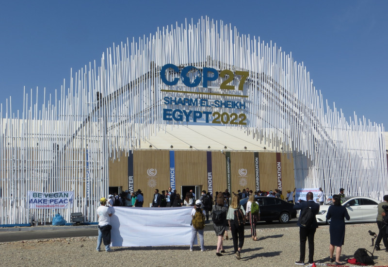 Gebäudeeingang, über dem steht: COP27, Sharm E--Sheikh Egypt2022. Davor Menschen, zum Teil  mit Transparenten oder Fotoapparaten.