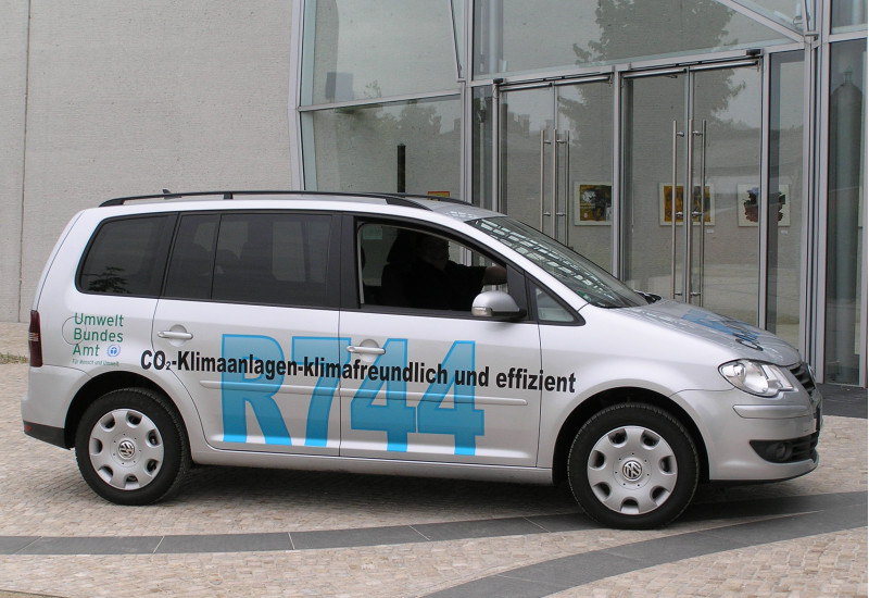 ein silbergrauer VW Touran steht vor dem UBA Dessau-Roßlau. Er trägt die Beschriftung "CO2-Klimaanlagen - klimafreundlich und effizient, R744" und das Logo des Umweltbundesamtes