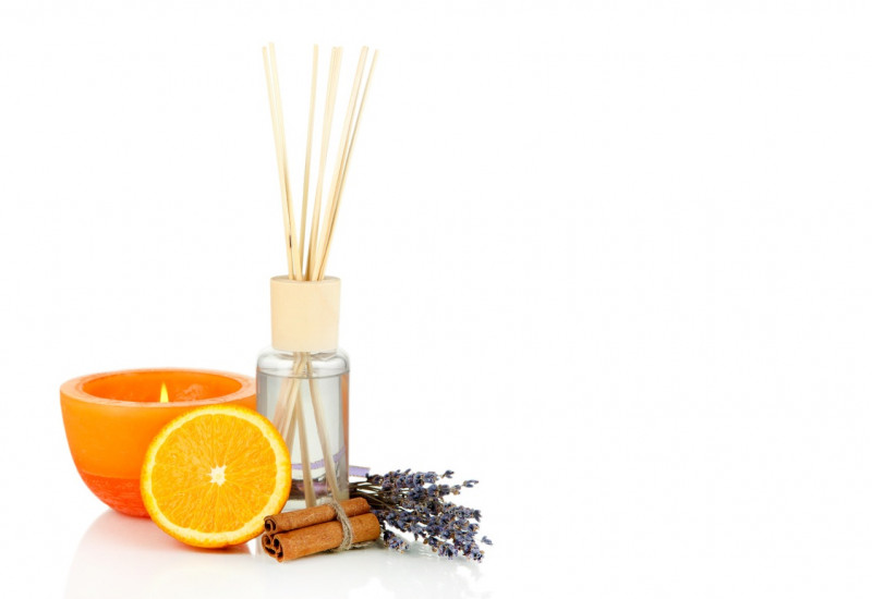 eine orange Duftkerze, ein Aromafläschchen mit Holzstäbchen zur Raumluft-Beduftung, eine halbe Orange, ein Lavendelsträußchen und Zimtstangen