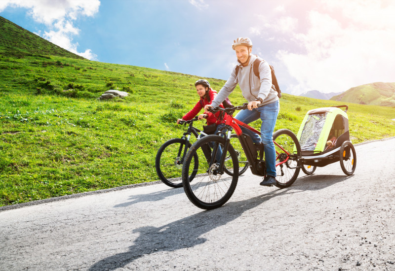 Mann auf einem E-Bike mit Kinder-Fahrradanhänger auf einer Straße in bergigem Gelände bei Sonnenschein