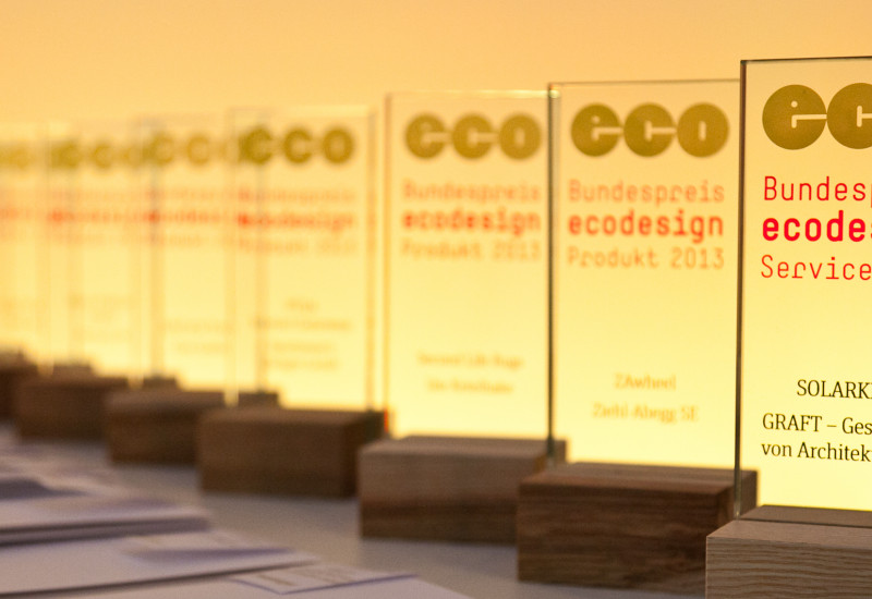 Auf einem Tisch stehen in einer Reihe die Sieger-Trophäen aus Glas mit dem Logo des Bundespreis Ecodesign und dem Namen des jeweiligen Preisträgers