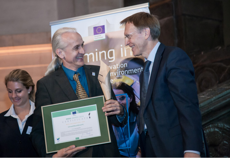 two smiling men, one is holding a certificate in his hands, in the background a poster with an EU flag