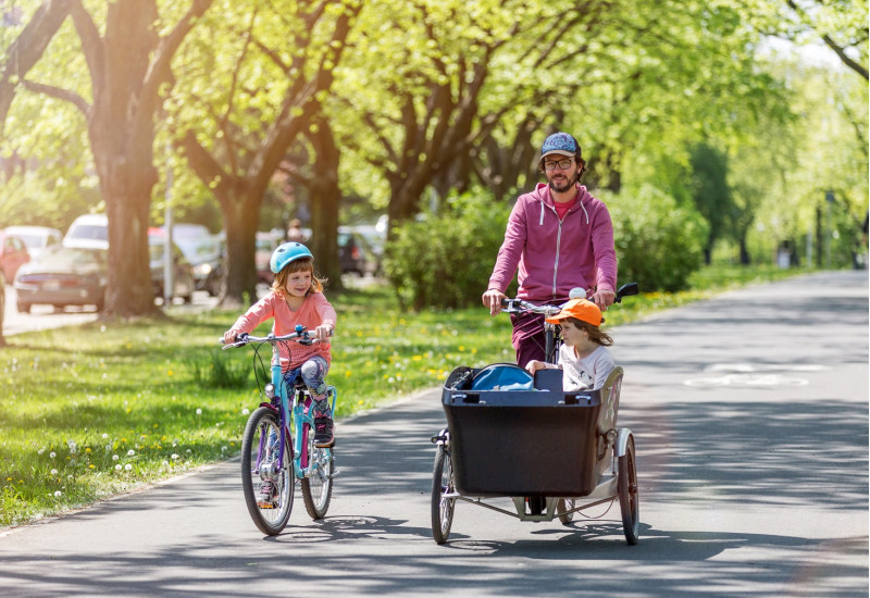breiter und begrünter Radweg in der Stadt, ein Vater transportiert seine kleine Tochter in einem Lastenrad, eine größere Tochter fährt mit eigenem Rad nebenher